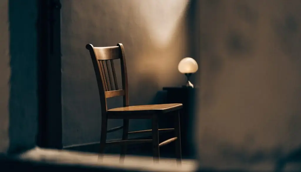 A single empty wooden chair beside a lit lamp in a quiet room.