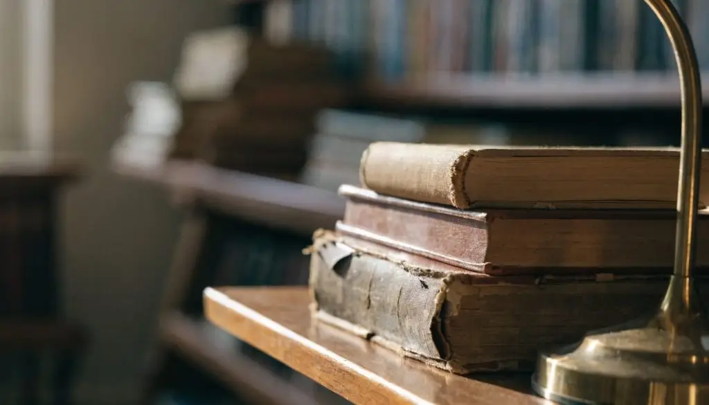 A stack of old cloth-bound books and reading glasses on a wooden desk in warm light.