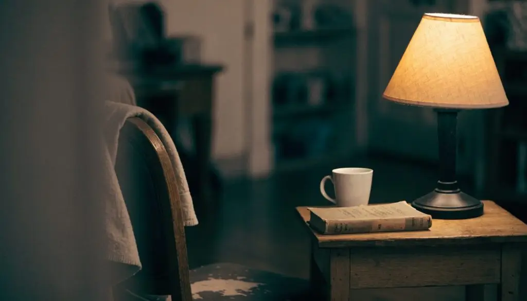 A single lit lamp beside an open Bible and a cup of coffee in a quiet room.