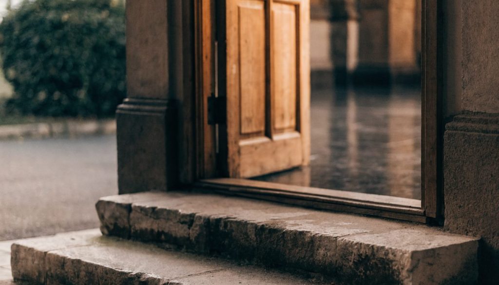 A plain wooden church door standing open with warm light spilling onto stone steps.