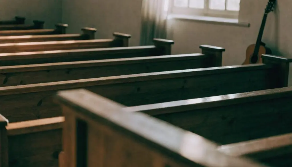 Empty wooden pews in soft morning light with an acoustic guitar leaning in the corner.