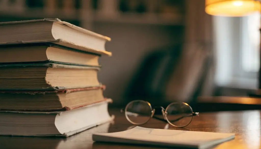 A stack of old cloth-bound books and reading glasses on a wooden desk in lamp light
