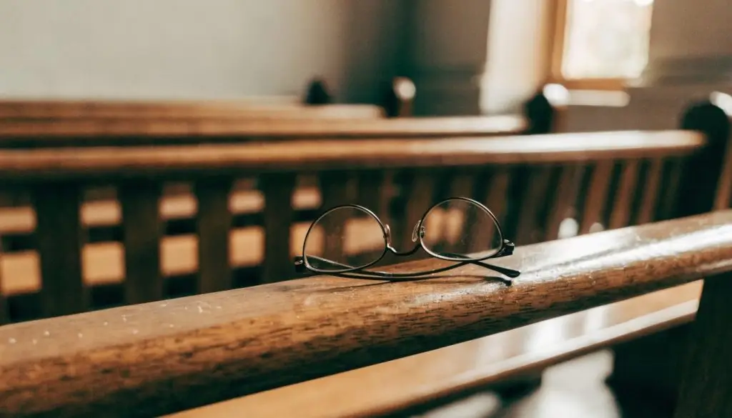 Old cloth-bound books and reading glasses resting on a wooden pew in soft window light